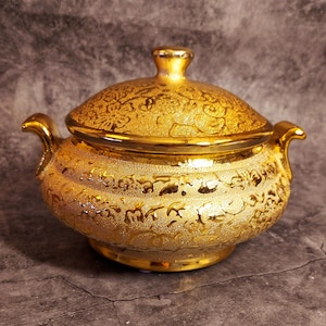 Könnte beinhalten: Eine goldene Keramik-Servierplatte mit Deckel. Die Platte ist mit einem komplizierten Blumenmuster verziert.
