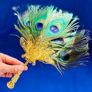 May include: A handheld fan featuring a cluster of iridescent green and blue peacock feathers arranged behind a decorative gold-toned metal filigree piece shaped like a lotus flower, attached to a long, ornate gold-toned metal handle.