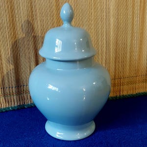 Może przedstawiać: Jasnoniebieski ceramiczny słoik na imbir z pasującą pokrywką. Słoik ma zaokrąglony korpus, podstawę i ozdobne zwieńczenie na pokrywce. Błyszczące wykończenie odbija światło. Odpowiedni do dekoracji wnętrz.