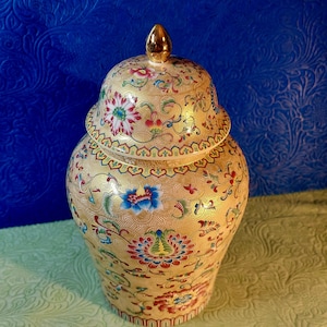 Peut inclure: Un pot en céramique décoratif avec un couvercle doré et des motifs floraux complexes dans des tons de rouge, bleu, vert et or sur un fond crème.