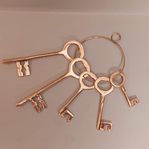 Może przedstawiać: Zestaw pięciu kluczy w antycznym stylu, wykonanych z mosiądzu, z pierścieniem przymocowanym do górnej części. Klucze są ułożone w kółko, z pierścieniem na górze.