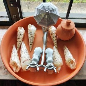 Puede incluir: Un plato de terracota con una escultura de plata en forma de paraguas en el centro. La escultura tiene una cadena que cuelga de ella y está rodeada de varias conchas. También hay una pequeña olla de terracota con tapa en el plato.