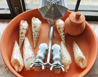 May include: A terracotta dish with a silver umbrella-shaped sculpture in the centre. The sculpture has a chain hanging from it and is surrounded by several seashells. There is also a small terracotta pot with a lid in the dish.