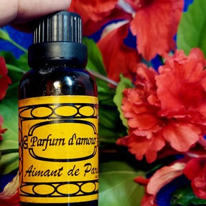 May include: A dark glass bottle with a black cap and a yellow label featuring the text "Parfum d'amour" and "Aimant de Paris". The bottle is held against a backdrop of red hibiscus flowers and green leaves.