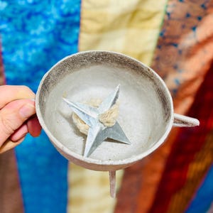 Könnte beinhalten: Ein silberfarbenes sternförmiges Objekt in einer kleinen, runden, silberfarbenen Schale mit Griff und kleinem Bein. Der Stern ruht auf einem beigefarbenen, faserigen Material. Der Hintergrund ist ein farbenfroher Patchwork aus blauen, gelben und roten Stoffen.