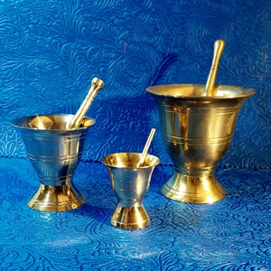 May include: Three brass mortar and pestle sets of varying sizes. The largest set has a pestle with a rounded end. The smallest set has a pestle with a flat end. The sets are on a blue patterned surface.