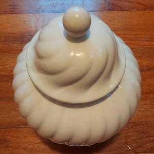 May include: A cream-coloured ceramic jar with a lid. The jar features a swirl design and a round knob on top of the lid. The jar is placed on a wooden surface. The jar is likely used for storing food items.