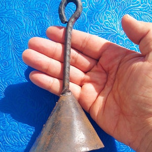 Puede incluir: Una campana de metal forjada a mano, con una base cónica y un asa en forma de lazo. La campana tiene un aspecto oscuro y envejecido, lo que sugiere que está hecha de hierro o un metal similar. La campana se sostiene en una mano sobre un fondo azul.