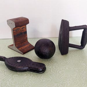 Op de afbeelding: Vier vintage deurstoppers van gietijzer in verschillende vormen, waaronder een spoorweg, een bal, een plat strijkijzer en een klein rond object met een handvat. De deurstoppers zijn allemaal zwart en hebben een rustieke, verweerde uitstraling.