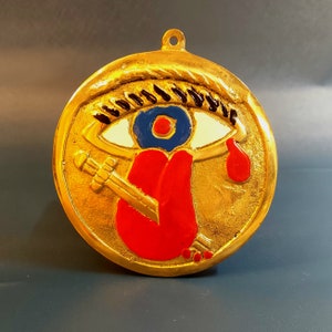 May include: A gold-coloured pendant with a stylized eye design. The eye features a blue iris, white and black accents, and a red teardrop. A red tongue and a sword are also depicted.