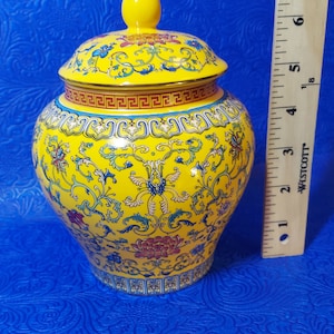 May include: A yellow ceramic jar with a lid, decorated with blue and red floral patterns. The jar has a gold trim around the top and bottom.