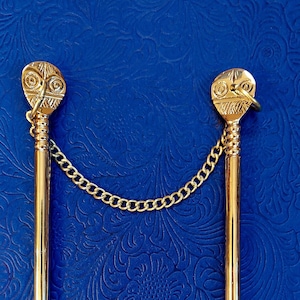May include: Two gold-colored hair sticks connected by a delicate chain. Each stick features a decorative head with an engraved design. The background is a textured, deep blue surface.