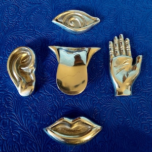 Könnte beinhalten: Fünf silberfarbene Metallfiguren in Form eines menschlichen Auges, Ohrs, einer Zunge, einer Hand und Lippen. Die Figuren sind auf einem blauen Hintergrund angeordnet.