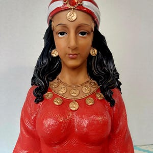 Op de afbeelding: Een vrouw met lang zwart haar draagt een rood-witte tulband met een rood juweel. Ze draagt een rode jurk met een gouden ketting en oorbellen. Ze houdt een rode kristallen bol in haar handen.