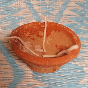 Puede incluir: Una pequeña lámpara de aceite de terracota sin esmaltar con forma de cuenco redondeado. La lámpara contiene un residuo de color claro y varias mechas de algodón blancas. La lámpara está sobre un fondo tejido azul y blanco.
