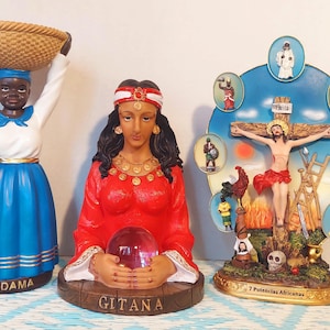 May include: Three religious figurines, one of a woman wearing a red dress and holding a crystal ball, one of a woman wearing a blue dress with a basket on her head, and one of a man on a cross with a circle of smaller figures around him. The figurines are all on wooden bases with text on them. The text on the bases reads "Gitana", "Madame", and "7 Potencias Africanas".