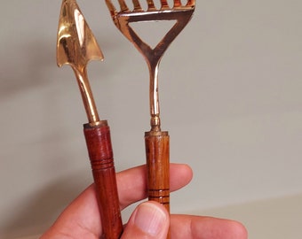 Peut inclure: Deux petits outils de jardinage avec des manches en bois. Les outils sont en laiton et ont une finition dorée. L'outil à gauche est une petite truelle et l'outil à droite est un petit râteau.