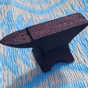 Pode incluir: Uma bigorna de metal preto com uma superfície texturizada. A bigorna está sobre uma superfície com um padrão azul e branco.