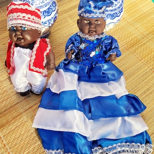 May include: Two dolls dressed in traditional clothing. The doll on the left is wearing a red and white outfit with a white hat. The doll on the right is wearing a blue and white dress with a blue hat.