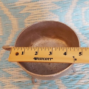 以下が含まれることがあります： 3本の脚が付いた小さな銀色の金属製の釜。釜の幅は約10cm、高さは約7.5cmです。青と白の模様の表面の上に置かれています。
