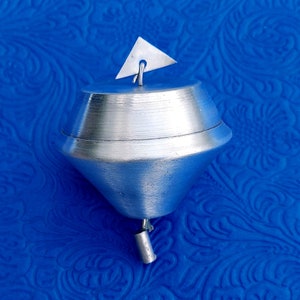 May include: A silver metal spinning top with a pointed top and a rounded base. The top is on a blue patterned surface.