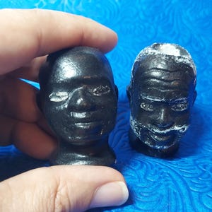 May include: Two black ceramic figurines of human heads. The heads are detailed with facial features and have a textured surface.