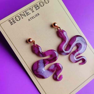 Può includere: Un paio di orecchini a forma di serpente viola e bianco con borchie color oro. Gli orecchini sono esposti su una carta beige con la scritta "HONEYBOO ATELIER". Lo sfondo è viola.