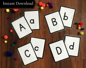 May include: A set of alphabet flashcards with black letters on white cards with black borders. The cards are scattered on a wood surface with colorful pom-poms.