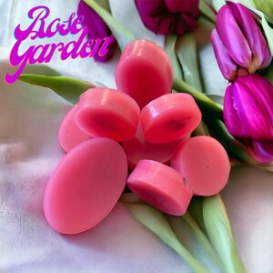 May include: Pink oval and round soaps are arranged on a white cloth with green leaves and purple tulips. The text "Rose Garden" is in pink script in the upper left corner. The soaps are various shades of pink.