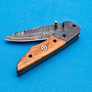 May include: A folding pocket knife with a wood handle and a patterned steel blade. The blade is partially open and the handle has a small red and gold design.