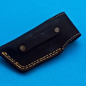 May include: A black leather knife sheath with a brown leather strap and two brass rivets. The sheath has a yellow stitching detail around the edges.