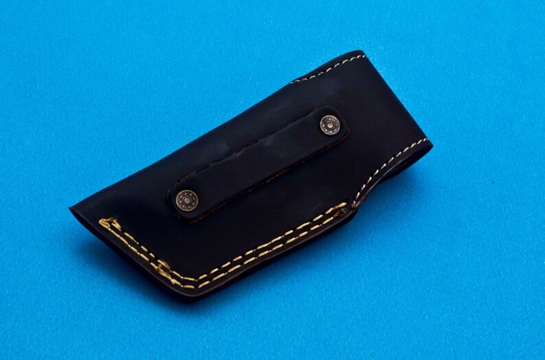 May include: A black leather knife sheath with a brown leather strap and two brass rivets. The sheath has a yellow stitching detail around the edges.
