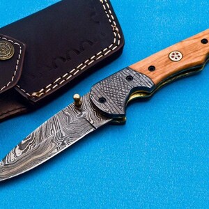 May include: A folding pocket knife with a wood handle and a patterned steel blade. The knife is closed and has a leather sheath.