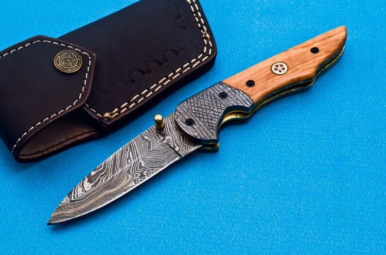 May include: A folding pocket knife with a wood handle and a patterned steel blade. The knife is closed and has a leather sheath.