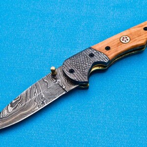 May include: A folding pocket knife with a wood handle and a patterned steel blade. The handle is brown with a red and white design. The blade is silver with a wavy pattern.