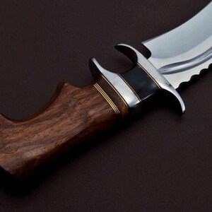 May include: A close-up of a hunting knife with a brown wooden handle and a silver blade. The blade has a serrated edge and a black and silver accent near the handle.