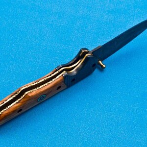 May include: A folding knife with a wooden handle and a black blade. The handle has a gold inlay design.