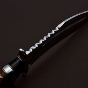 May include: A close-up of a hunting knife with a wooden handle and a serrated blade. The blade is made of stainless steel and has a black finish.