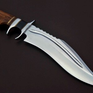 May include: A large, silver, curved knife with a wooden handle. The handle has a black and gold inlay. The blade has a serrated edge and a single fuller.