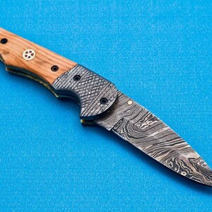 May include: A folding knife with a wooden handle and a patterned steel blade. The blade is open and the knife is lying on a blue surface.