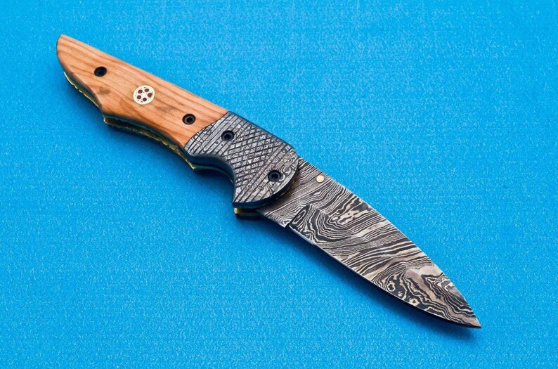 May include: A folding knife with a wooden handle and a patterned steel blade. The blade is open and the knife is lying on a blue surface.