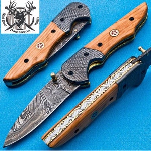May include: A folding pocket knife with a Damascus steel blade and a wooden handle. The handle has a decorative inlay of gold and silver. The knife is closed and the blade is visible. The knife is made by Wild Deer Damascus Knife.