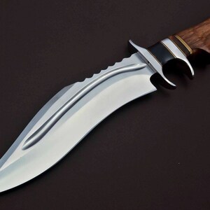 May include: A silver-colored hunting knife with a brown wooden handle. The blade has a serrated edge and a curved shape.
