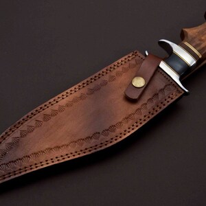 May include: A brown leather sheath with decorative stitching and a brass button. A hunting knife with a wooden handle is inside the sheath.