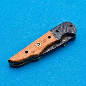 May include: A folding knife with a wooden handle and a patterned steel blade. The handle has a red and gold inlay.