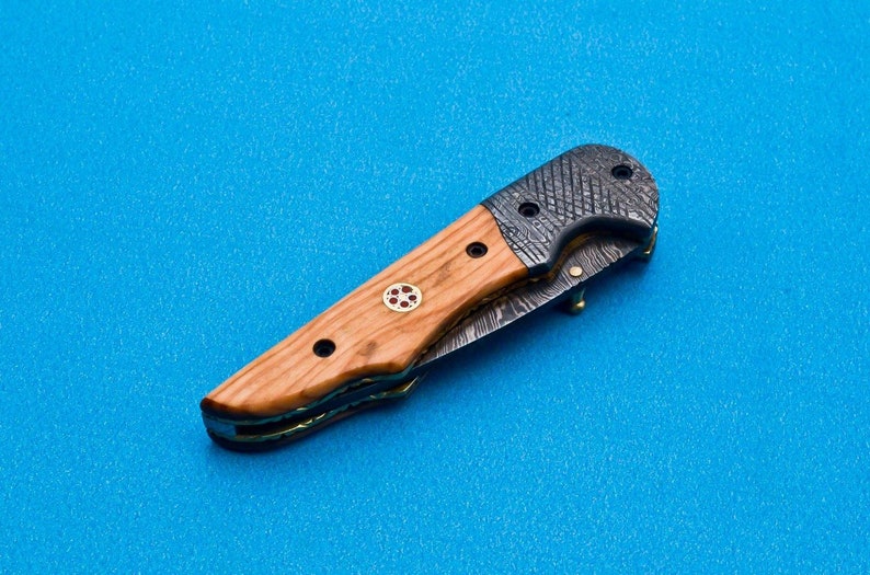 May include: A folding knife with a wooden handle and a patterned steel blade. The handle has a red and gold inlay.