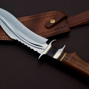 May include: A curved, silver-colored knife with a wooden handle and a leather sheath. The knife has a serrated edge and a decorative metal band around the handle.