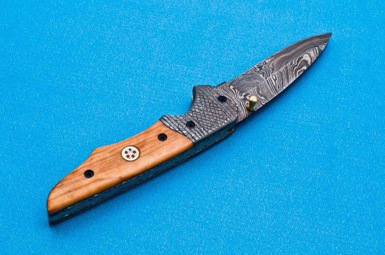 May include: A folding knife with a wooden handle and a patterned steel blade. The handle has a red and white design.