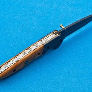 May include: A black folding knife with a wooden handle and a silver and gold inlay design. The blade is open and the knife is lying on a blue surface.