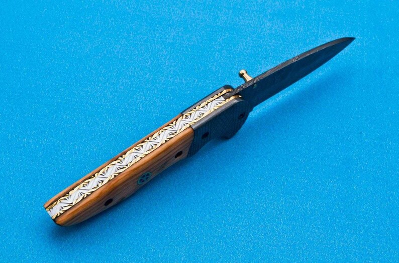 May include: A black folding knife with a wooden handle and a silver and gold inlay design. The blade is open and the knife is lying on a blue surface.